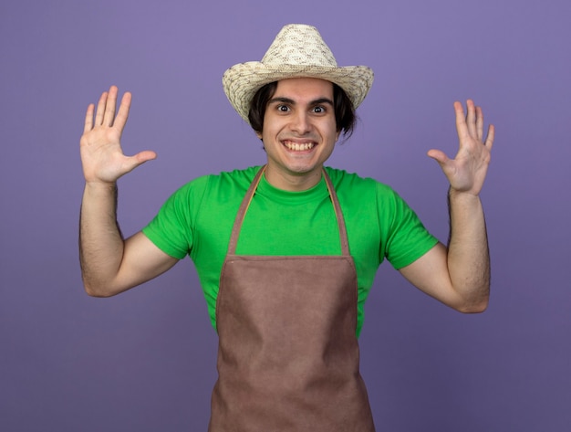 Jardinero macho joven sonriente en uniforme vistiendo sombrero de jardinería levantando las manos