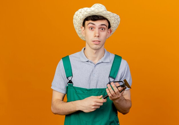 Jardinero macho joven sorprendido con sombrero de jardinería tiene rastrillo de azada