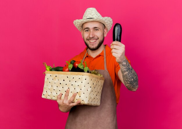 Jardinero macho sonriente vistiendo sombrero de jardinería tiene canasta de verduras y berenjena