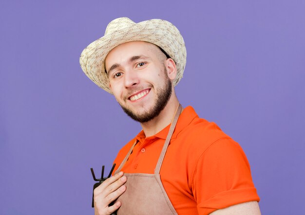 Jardinero macho sonriente vistiendo sombrero de jardinería tiene rastrillo azada