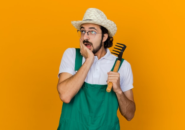 Jardinero macho sorprendido en gafas ópticas con sombrero de jardinería pone la mano en la barbilla y sostiene el rastrillo mirando al lado
