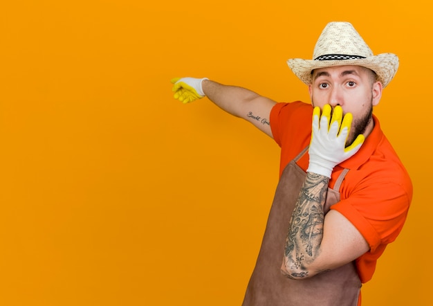 Foto gratuita jardinero macho sorprendido vistiendo guantes y gorro de jardinería pone la mano en la boca apuntando hacia atrás y mirando