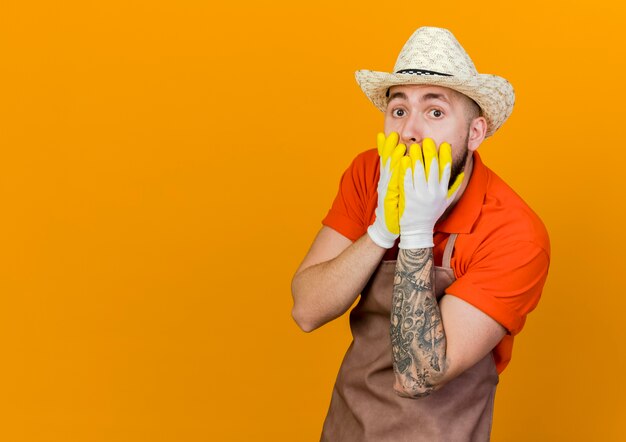 Jardinero macho sorprendido vistiendo guantes y gorro de jardinería pone las manos en la boca