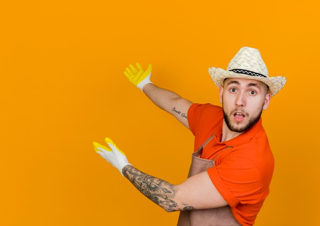 Jardinero macho sorprendido vistiendo guantes y sombrero de jardinería puntos en la espalda con dos manos mirando