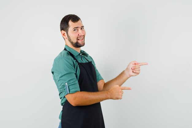 Jardinero masculino en camiseta, delantal apuntando hacia el lado y mirando alegre, vista frontal.