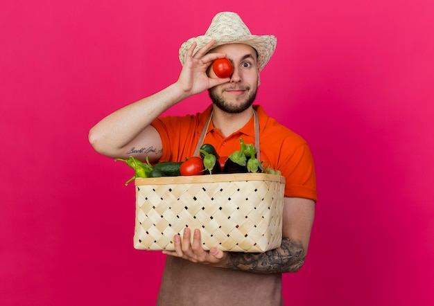 Jardinero masculino complacido vistiendo sombrero de jardinería tiene canasta de verduras