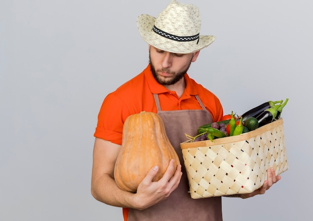 Jardinero masculino confiado con sombrero de jardinería sostiene canasta de verduras y mira calabaza