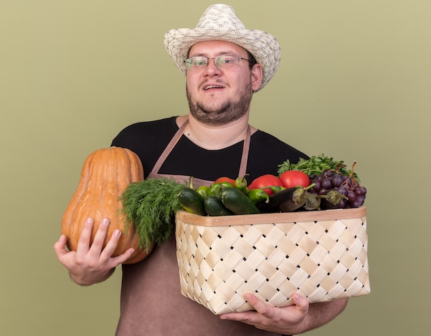 Jardinero masculino joven complacido con sombrero de jardinería sosteniendo canasta de verduras con calabaza aislado en la pared verde oliva