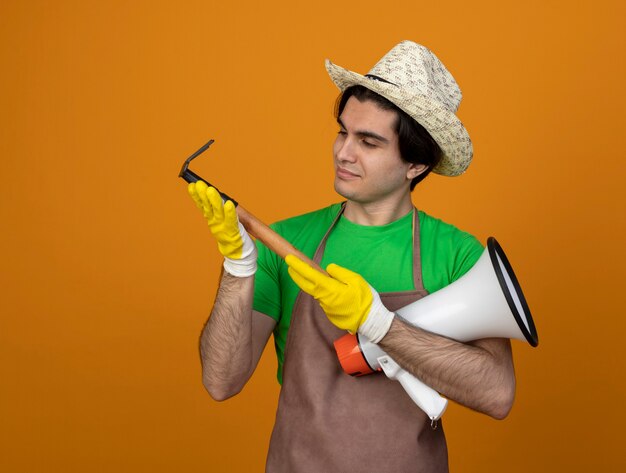 Jardinero masculino joven complacido en uniforme con sombrero de jardinería con guantes sosteniendo altavoz y mirando rastrillo en su mano aislado en naranja