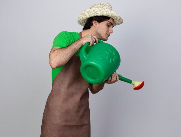 Jardinero masculino joven complacido en uniforme vistiendo sombrero de jardinería regar con regadera