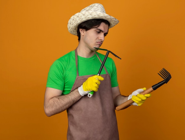 Jardinero masculino joven disgustado en uniforme con sombrero de jardinería con guantes sosteniendo y mirando azada rastrillo con rastrillo aislado en naranja