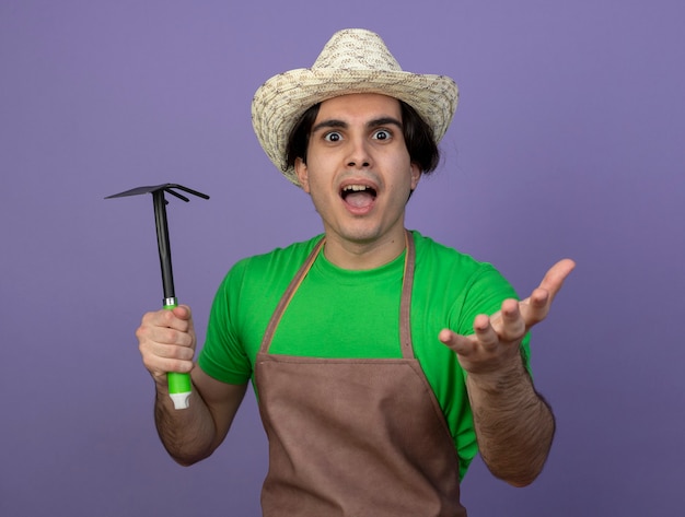 Jardinero masculino joven emocionado en uniforme con sombrero de jardinería sosteniendo rastrillo de azada aislado en púrpura