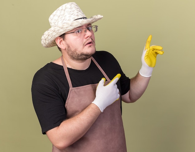 Jardinero masculino joven impresionado con sombrero de jardinería y guantes puntos con los dedos detrás aislado en la pared verde oliva con espacio de copia