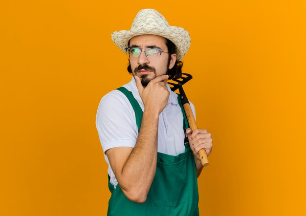 Jardinero masculino pensativo en gafas ópticas con sombrero de jardinería pone la mano en la barbilla y sostiene el rastrillo mirando al lado