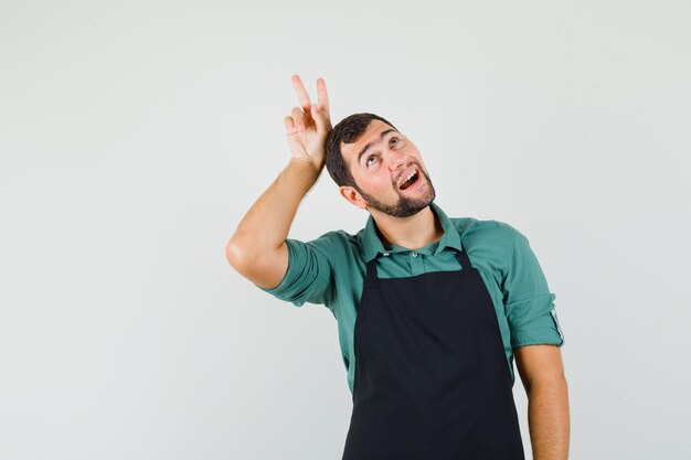 Jardinero masculino que muestra el signo v detrás de la cabeza como cuernos en una camiseta, delantal y con un aspecto divertido, vista frontal.