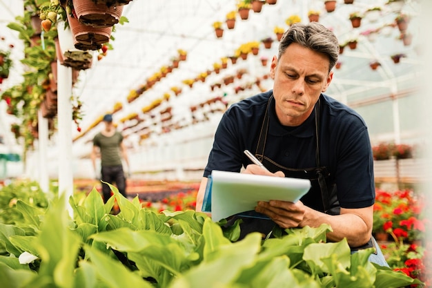 Jardinero masculino revisando las existencias en el vivero de plantas y tomando notas en un portapapeles