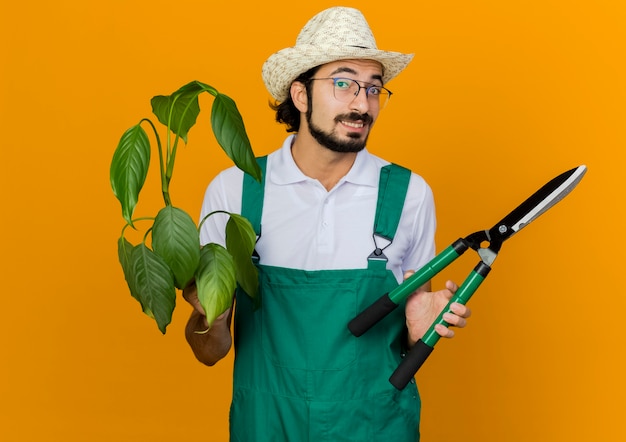 Jardinero masculino sonriente en gafas ópticas con sombrero de jardinería tiene podadoras y plantas