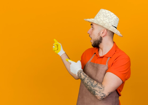 Jardinero masculino sorprendido con sombrero y guantes de jardinería mira y apunta hacia atrás con las dos manos