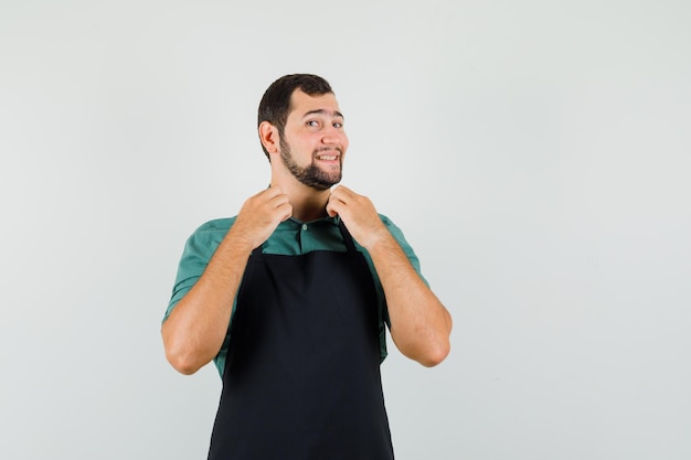 Jardinero masculino sosteniendo su cuello en camiseta, delantal y mirando alegre, vista frontal.