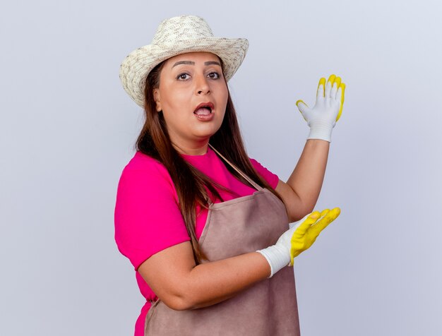 Jardinero de mediana edad mujer en delantal y sombrero con guantes de goma presentando algo con los brazos mirando a la cámara sorprendido de pie sobre fondo blanco.