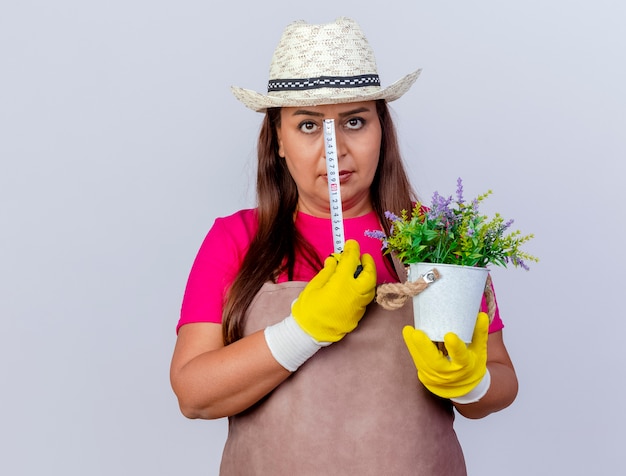Foto gratuita jardinero de mediana edad mujer en delantal y sombrero con guantes de goma sosteniendo planta en maceta y cinta métrica mirando a cámara con rostro serio de pie sobre fondo blanco