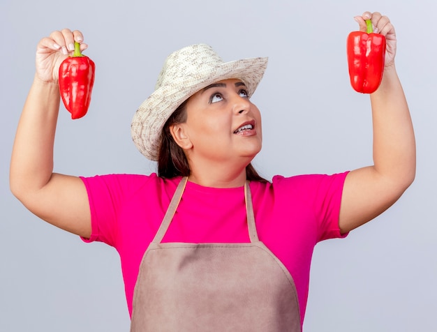 Jardinero de mediana edad mujer en delantal y sombrero mostrando pimientos rojos frescos sonriendo mirándolos de pie sobre fondo blanco.