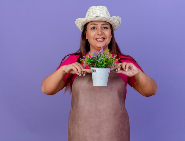 Jardinero de mediana edad mujer en delantal y sombrero mostrando planta en maceta mirando a cámara sonriendo con cara feliz de pie sobre fondo púrpura