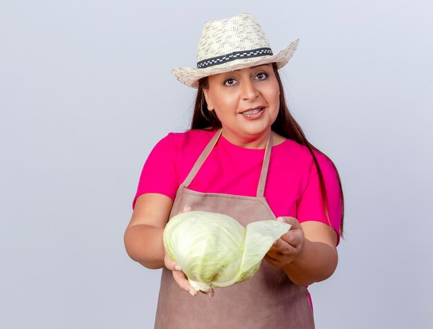 Jardinero de mediana edad mujer en delantal y sombrero mostrando repollo sonriendo