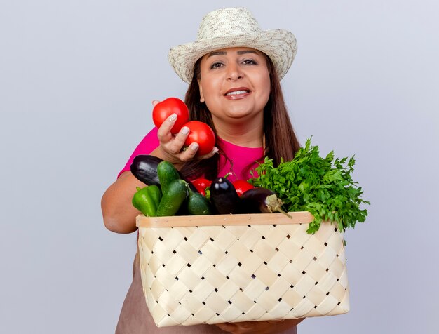 Jardinero de mediana edad mujer en delantal y sombrero sosteniendo cajón lleno de verduras sonriendo con cara feliz
