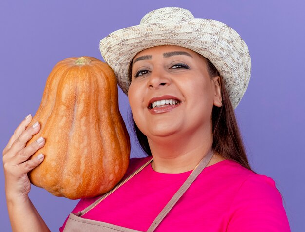 Jardinero de mediana edad mujer en delantal y sombrero sosteniendo calabaza con sonrisa en la cara