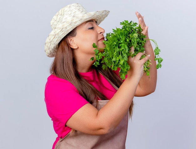 Jardinero de mediana edad mujer en delantal y sombrero sosteniendo hierbas frescas inhalando buen aroma de pie sobre fondo blanco.