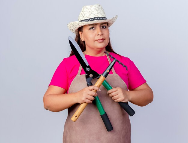 Jardinero de mediana edad mujer en delantal y sombrero sosteniendo mini rastrillo y cortasetos mirando a cámara con cara seria de pie sobre fondo blanco.