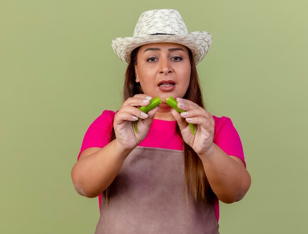 Jardinero de mediana edad mujer en delantal y sombrero sosteniendo mitades de ají verde mirando confundido parado sobre fondo claro