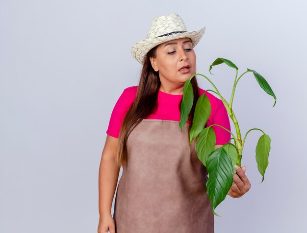 Jardinero de mediana edad mujer en delantal y sombrero sosteniendo la planta mirando intrigado