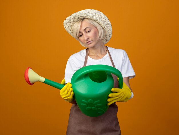 Jardinero de mediana edad mujer en uniforme de jardinero con sombrero y guantes de jardinería sosteniendo y mirando regadera