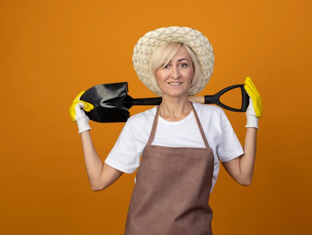 Jardinero de mediana edad sonriente mujer en uniforme de jardinero con sombrero y guantes de jardinería sosteniendo la pala detrás del cuello mirando a la cámara aislada sobre fondo naranja