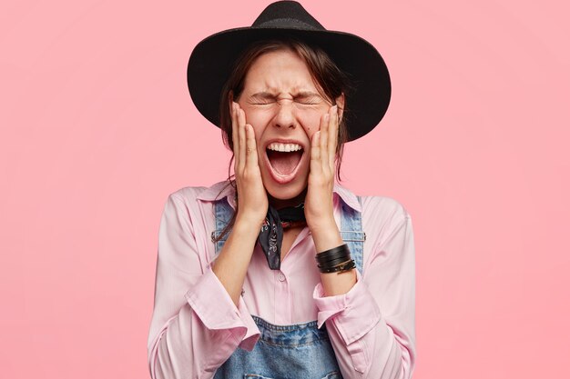 Jardinero mujer atractiva mantiene los ojos cerrados, abre la boca ampliamente, toca las mejillas, usa sombrero y overoles