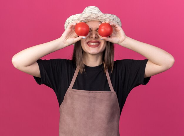 Jardinero mujer bastante caucásica ansioso vistiendo sombrero de jardinería sosteniendo tomates delante de los ojos