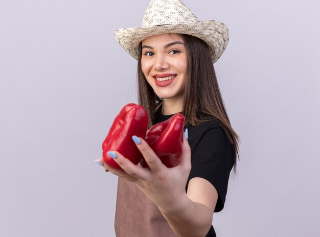 Jardinero mujer bonita caucásica sonriente vistiendo sombrero de jardinería sosteniendo pimientos rojos aislado en la pared blanca con espacio de copia