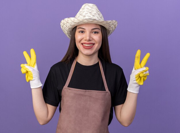 Jardinero mujer caucásica bastante sonriente vistiendo guantes y sombrero de jardinería gesticulando el signo de la victoria