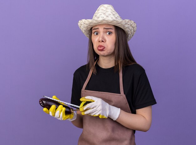 Jardinero mujer caucásica bastante triste vistiendo gorro de jardinería y guantes de medición de berenjena con cinta métrica