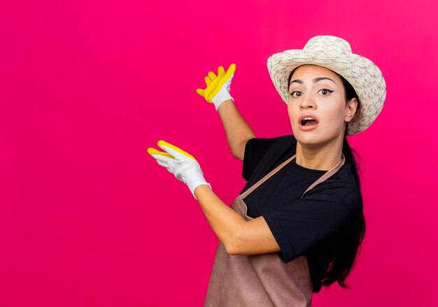 Jardinero de mujer hermosa joven en delantal de guantes de goma y sombrero presentando algo hacia atrás con los brazos sobre la pared rosa