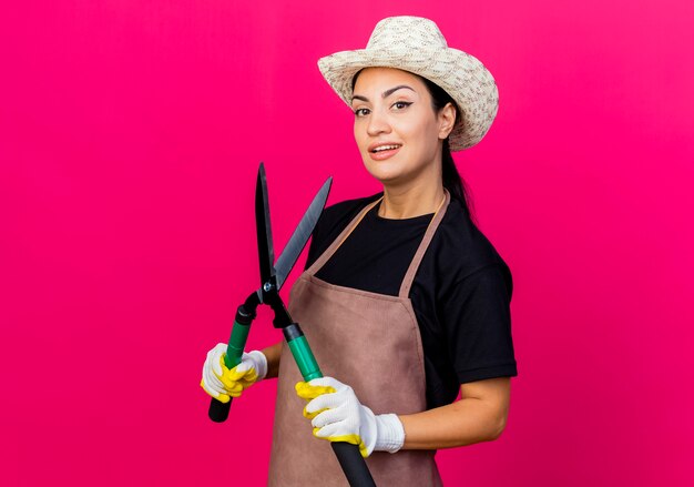 Jardinero de mujer hermosa joven en delantal de guantes de goma y sombrero sosteniendo tijeras de podar mirando al frente sonriendo con cara feliz de pie sobre la pared rosa