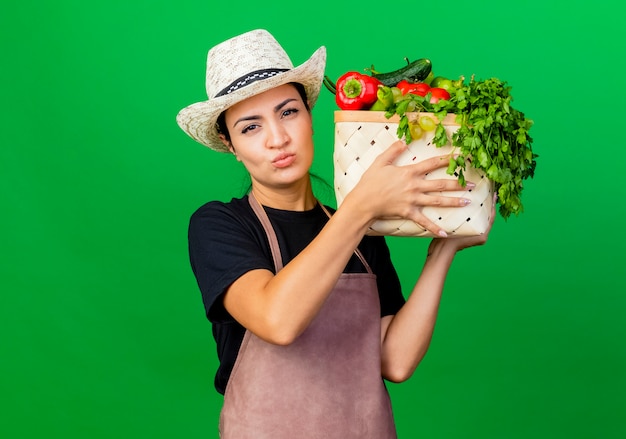 Jardinero de mujer hermosa joven en delantal y sombrero con caja llena de verduras con cara seria