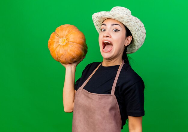 Jardinero de mujer hermosa joven en delantal y sombrero con calabaza feliz y emocionado