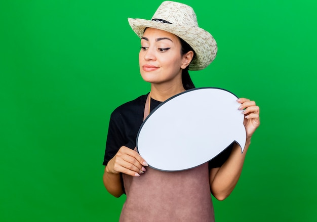 Jardinero de mujer hermosa joven en delantal y sombrero con cartel de burbujas de discurso en blanco mirando a un lado con una sonrisa en la cara de pie sobre la pared verde