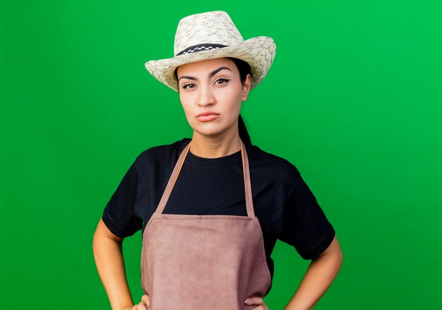 Jardinero de mujer hermosa joven en delantal y sombrero con expresión escéptica seria