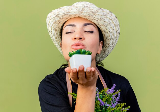 Jardinero de mujer hermosa joven en delantal y sombrero mostrando plantas en macetas que sopla un beso de pie sobre la pared verde claro