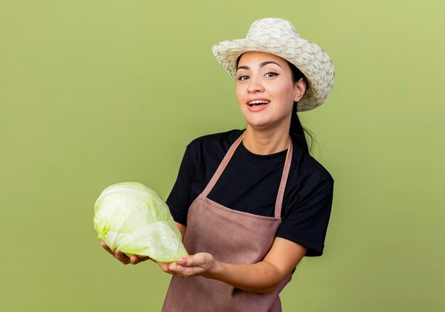 Jardinero de mujer hermosa joven en delantal y sombrero mostrando repollo mirando al frente sonriendo de pie sobre la pared verde claro