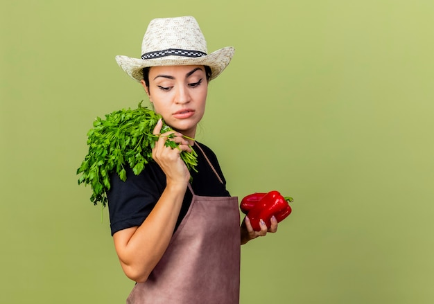 Jardinero de mujer hermosa joven en delantal y sombrero con pimiento rojo y hierbas frescas mirando a un lado con cara seria de pie sobre la pared verde claro